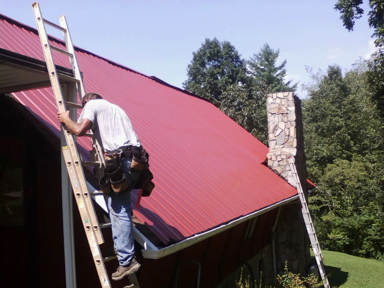Roofer on a ladder beside a red metal roof and stone chimney — Soffit & Fascia Repair by Roofing Brothers in Eckley